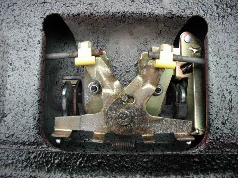 1990 Ford F-250 - Close-up of a metal latch mechanism with springs, rods and yellow plastic clips inside a recessed sheet-metal cavity.