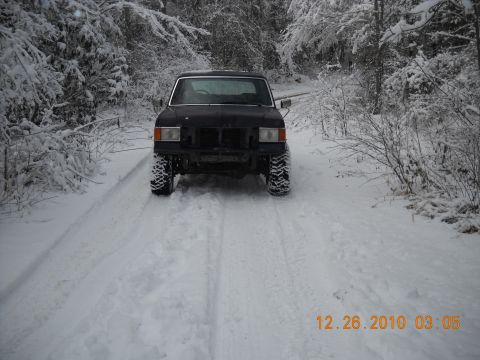1990 Ford Bronco - A black SUV drives down a snow-covered road surrounded by trees.
