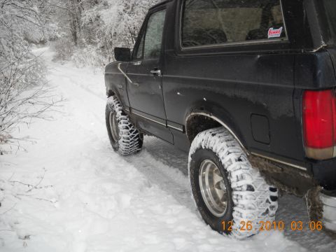 1990 Ford Bronco - A black Ford Bronco drives down a snow-covered road in a wooded area.