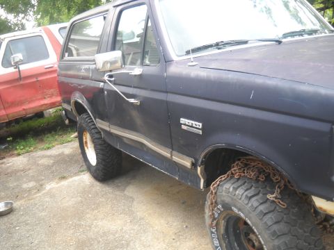 1990 Ford Bronco - A dark blue Ford Bronco sits next to a red and white truck, showing chains on its front tire.