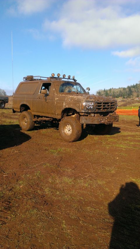 1995 Ford Bronco - This heavily mud-covered Ford Bronco, likely from the early 1990s, navigates a farm field under a bright blue sky.