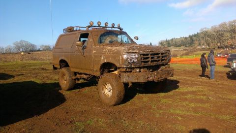 1995 Ford Bronco - A heavily mud-caked SUV sits in a muddy outdoor area, with trees and a clear sky in the background.