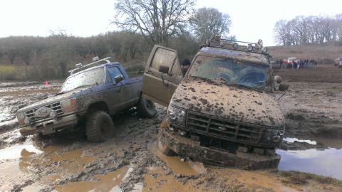 1995 Ford Bronco - Two off-road vehicles are deeply mired in thick mud, with one truck's door open and a person visible inside.