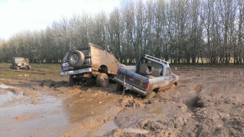 1995 Ford Bronco - Two trucks are stuck in deep mud on a farm, with trees in the background.