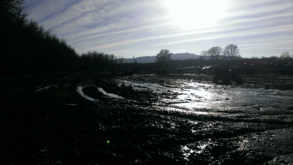 Muddy field with tire tracks and several dark vehicles in the distance under a cloudy sky