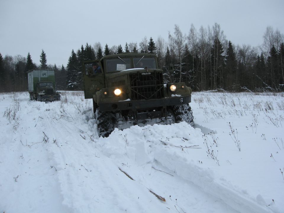 Green military truck driving through deep snow, another truck visible in background