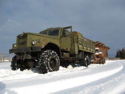 1987 Ford Bronco - A large, olive green military truck with aggressive tires navigates a snowy landscape, parked beside a tractor and a modern wooden building.