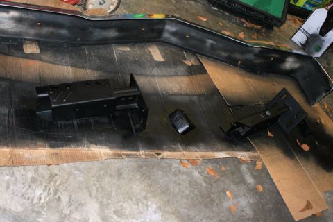 1976 Ford Ranchero - Black car bumper and mounting brackets are laid out on a surface covered with cardboard and wood planks.