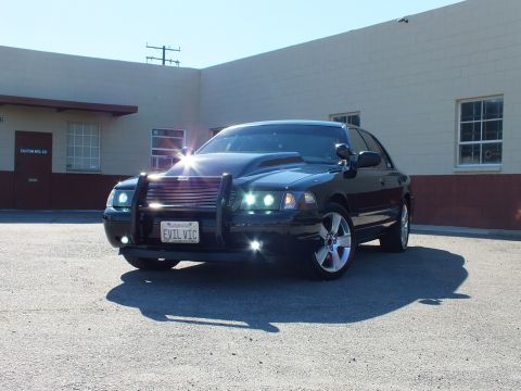 2003 Ford Crown Victoria - A black Ford Crown Victoria is parked in front of a commercial building, a push bar and a hood scoop.
