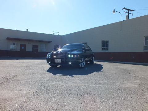 2003 Ford Crown Victoria - Distant front view of a custom black Crown Victoria, headlights on, beneath utility poles and clear sky.