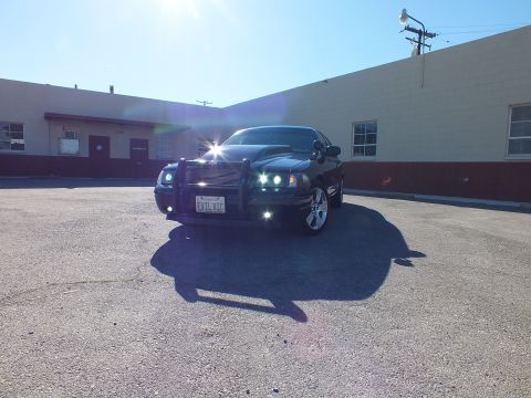 2003 Ford Crown Victoria - Wide front shot of a black Crown Victoria with grille guard against tan warehouse under bright sun.