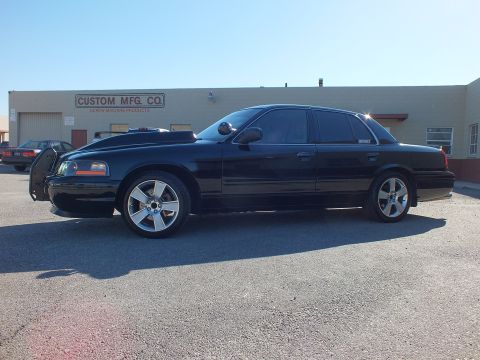 2003 Ford Crown Victoria - Driver-side view of a black Crown Victoria sedan, brush guard up front, parked along a warehouse.