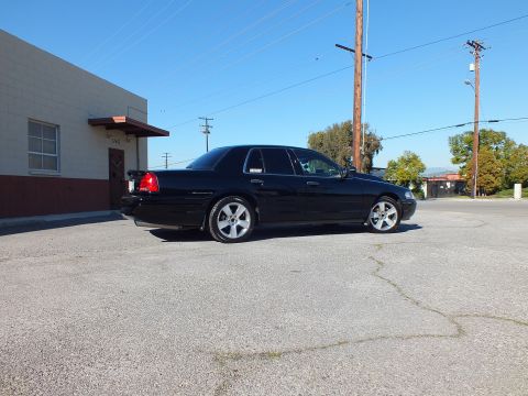 2003 Ford Crown Victoria - Black sedan photographed from rear quarter, deck spoiler and chrome rims, on weathered asphalt