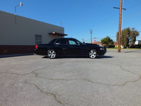 2003 Ford Crown Victoria - Profile of a black full-size sedan with chrome rims and trunk spoiler in a parking lot under clear sky
