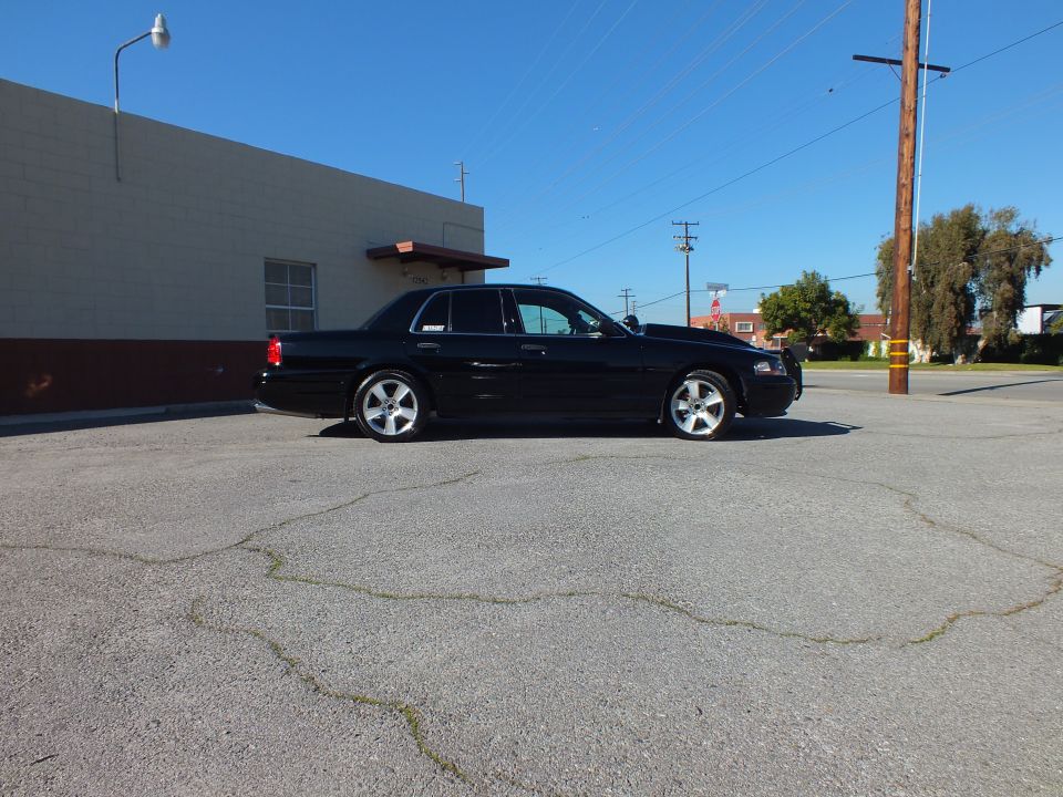 Black Crown Victoria sedan driver-side profile with chrome wheels parked beside gray building