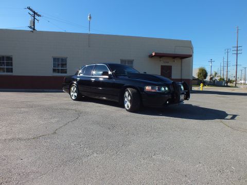 2003 Ford Crown Victoria - Black sedan with push-bar grille guard and quad projector lights parked beside a beige building