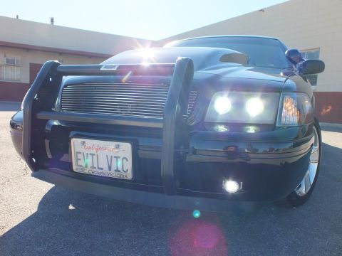 2003 Ford Crown Victoria - Low front-quarter view of a black sedan with grille guard and billet grille, EVIL VIC plate lit by sun