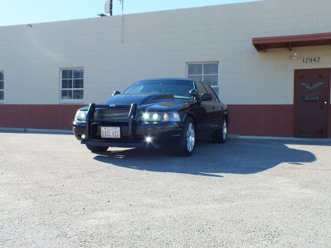 2003 Ford Crown Victoria - Black sedan shown from front quarter, grille guard and chrome rims, building number 12942 behind