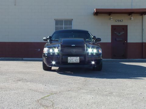 2003 Ford Crown Victoria - Straight-on black sedan with grille guard and lit lights, EVIL VIC plate, parked at building 12942