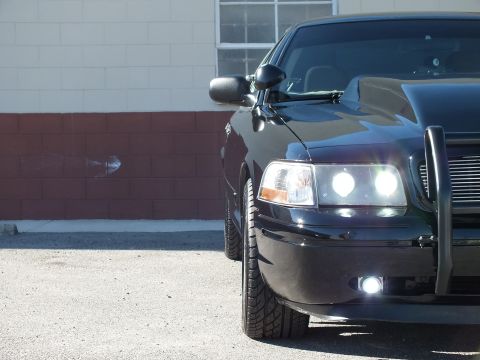2003 Ford Crown Victoria - Close front-quarter view of a black sedan, headlights and fog lamps lit, black push bumper against a brick wall.