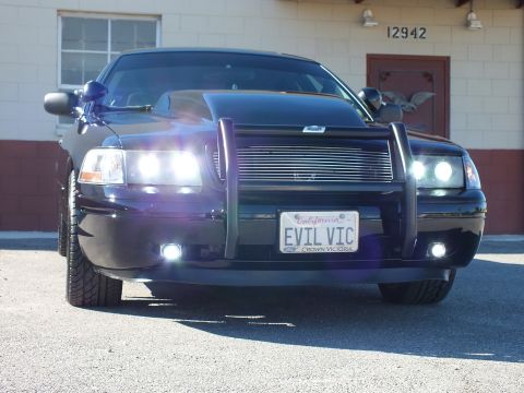 2003 Ford Crown Victoria - Black sedan photographed head-on, headlights glowing, EVIL VIC plate below a chrome billet grille.