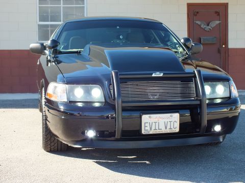 2003 Ford Crown Victoria - Black sedan front end with dual projector headlights, billet grille and the EVIL VIC plate, brick wall behind.