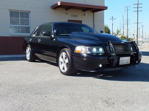 2003 Ford Crown Victoria - Lowered black sedan at a front-quarter angle, EVIL VIC plate and aftermarket wheels, building in background.