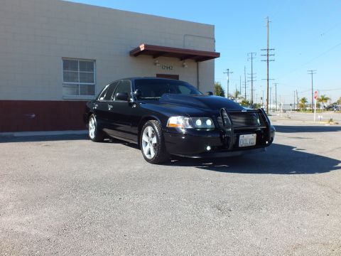 2003 Ford Crown Victoria - Black sedan shot from the front quarter on cracked asphalt, push bar fitted, stop sign and poles behind.