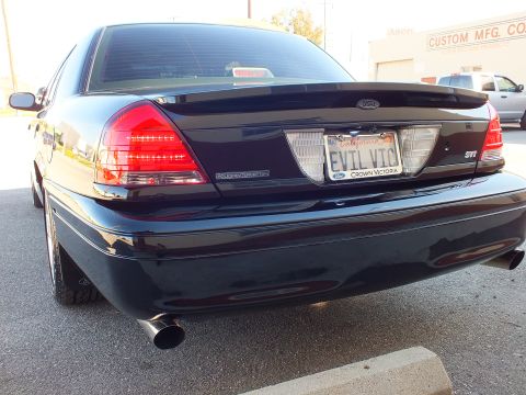 2003 Ford Crown Victoria - Black sedan rear shot with EVIL VIC plate and SVT badge, dual exhaust, a Custom Mfg sign in the background.