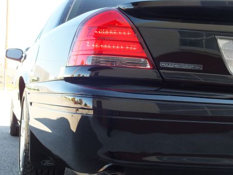 2003 Ford Crown Victoria - Close rear-corner detail of a black sedan showing a Police Interceptor badge and a Ford wheel emblem.