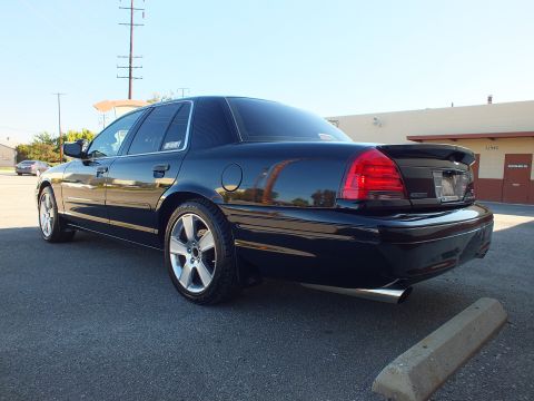 2003 Ford Crown Victoria - Driver-side rear-quarter view of a black lowered sedan on polished wheels parked beside a building.