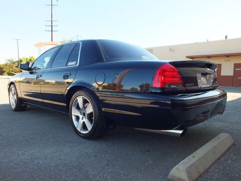 2003 Ford Crown Victoria - Black four-door sedan photographed from rear left, dual exhaust tips and tinted windows visible against industrial building.