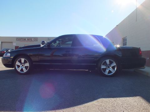 2003 Ford Crown Victoria - Full side profile of a lowered black sedan on polished wheels, sun glare across the body, warehouse behind.