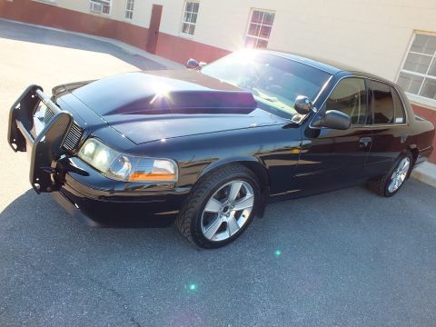 2003 Ford Crown Victoria - Black sedan angled front view showing grille guard, raised hood scoop, projector lights and polished wheels.