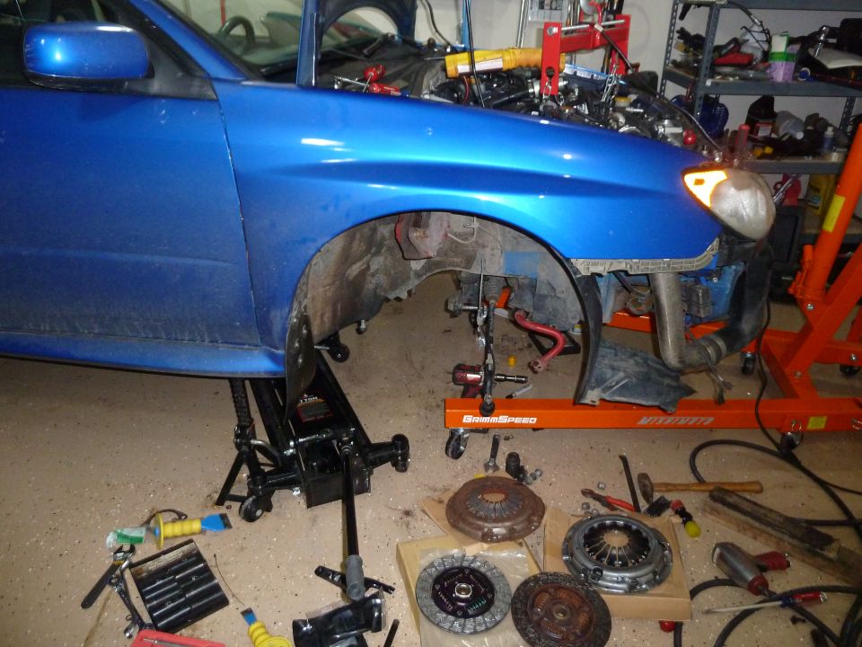 Blue Subaru Impreza with front end disassembled, engine exposed, and clutch parts laid out on the floor