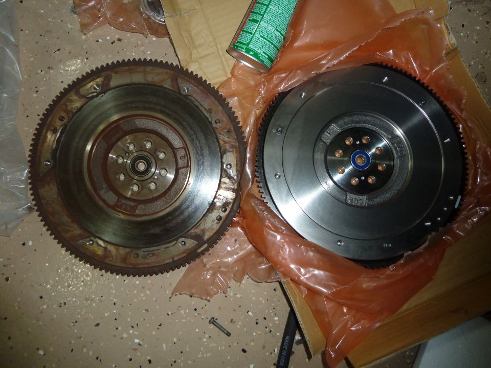 Two flywheels, one old and rusty, the other new and shiny, sit on a garage floor