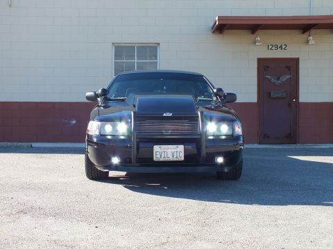 2003 Ford Crown Victoria - Symmetric front view of a black sedan, all lights lit, brush guard and EVIL VIC plate before a wall marked 12942.