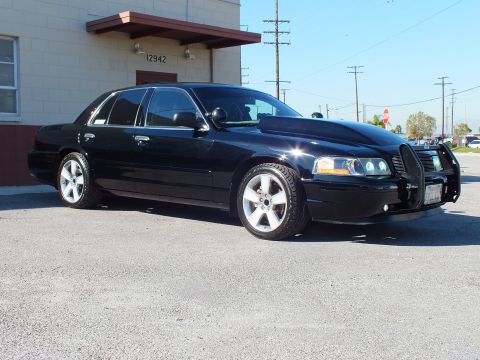 2003 Ford Crown Victoria - Front right view of a black sedan on polished wheels, brush guard and billet grille, parked by a warehouse.