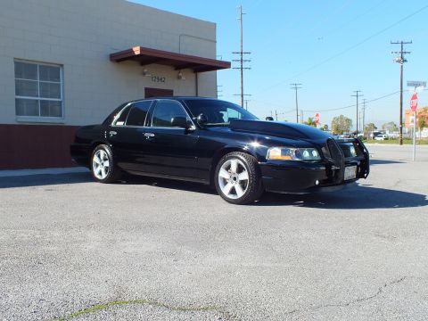 2003 Ford Crown Victoria - Black four-door sedan from front right at a distance, brush guard and polished wheels, blue sky overhead.