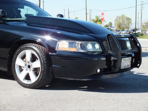 2003 Ford Crown Victoria - Detail of a black sedan's front corner showing projector headlight, tubular guard edge and polished five-spoke wheel.