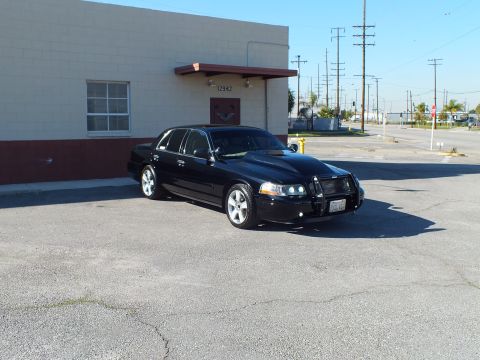 2003 Ford Crown Victoria - Distant front-right view of a black sedan with grille guard and projector headlights in an industrial lot.