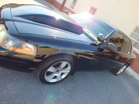 2003 Ford Crown Victoria - Low-angle shot of a black sedan's hood, mirror and machined alloy wheel beside a brick wall with white windows