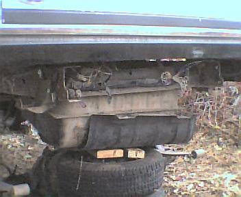 1984 Ford Bronco - Underside view of a rear axle and fuel tank assembly, supported by tires and wood blocks. The SuperMotors logo is visible at the bottom rig…