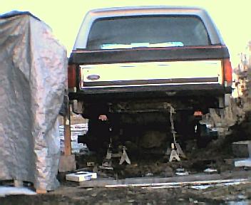 1984 Ford Bronco - The rear of a Ford Bronco is elevated on jack stands, undergoing work, with a tarp covering something to the left.