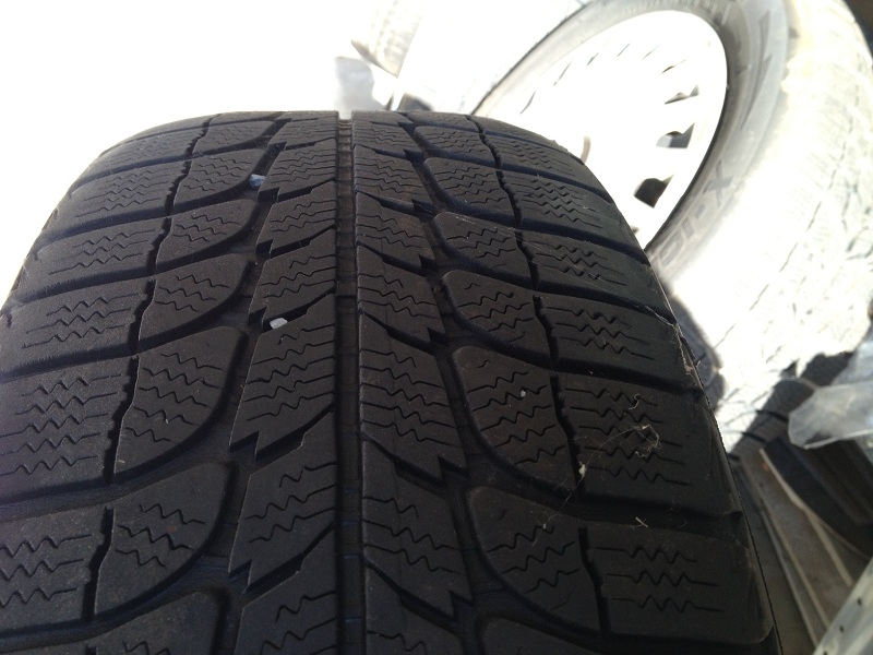 Close-up of a tire tread with a small object embedded in it. A white wheel is visible in the background