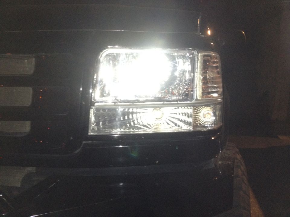 Close-up of a vehicle's headlight assembly with the light on. Watermark reads SUPERMOTORS