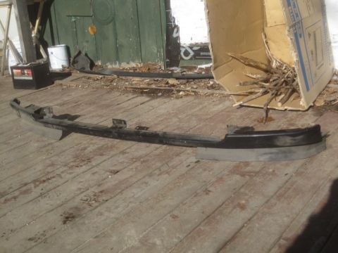 1993 Ford Bronco - A black front bumper part is displayed on weathered wooden planks, with a cardboard box and debris in the background.