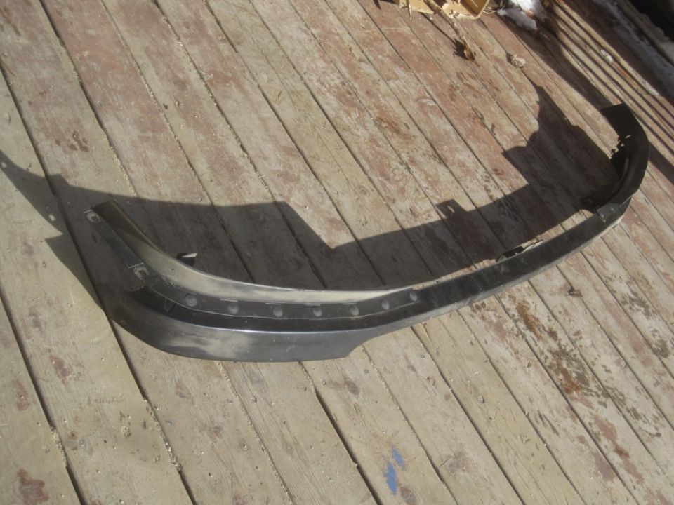 Black plastic bumper cover with small bolt holes along the top edge, lying on wooden planks