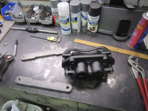 2008 Jeep Wrangler - A black brake caliper and two metal mounting brackets are laid out on a workbench with various tools and spray cans.