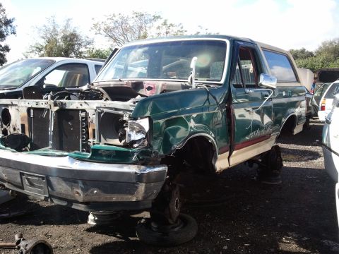 1994 Ford Bronco - A damaged green Ford Bronco sits in a junkyard, missing its hood and grille.